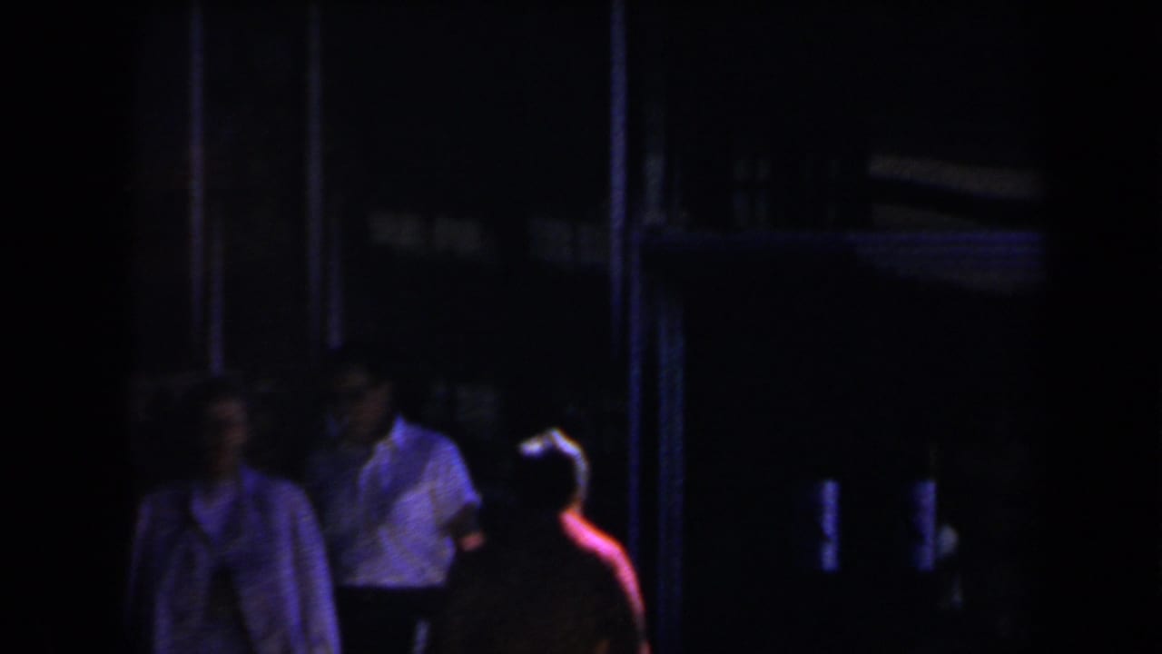 Two shadowed people near a dark building entrance, one wearing red and another in a light shirt.