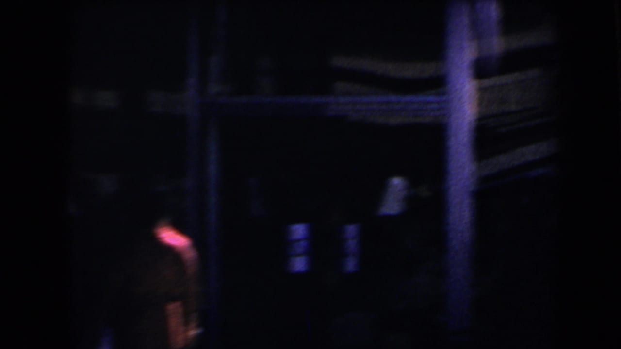 Dark nighttime street view with blurred figure, building shadows, and small lit windows.
