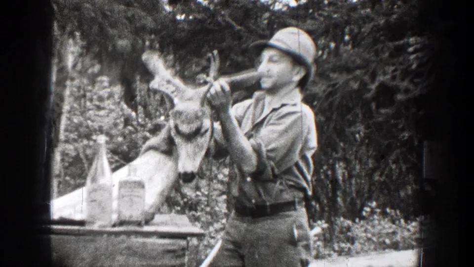 Chelan, Washington 1939 frame showing a man in a hat holding or presenting a deer skull with antlers beside bottles on an outdoor table.