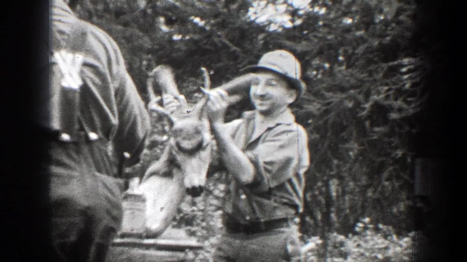 A 1939 Chelan home-movie frame shows a smiling man in a hat presenting a deer skull with antlers, with bottles and another adult partly visible.