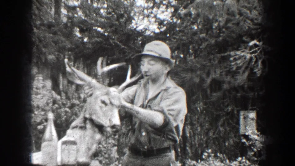 In 1939 Chelan, Washington home-movie footage, a man in a brimmed hat and button-front shirt holds a buck head with antlers beside bottles on an outdoor table.