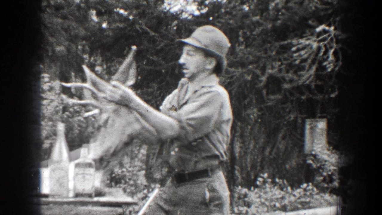 A man in late-1930s Chelan, Washington footage steadies a deer head with antlers near bottles on a table, with trees and brush behind him.
