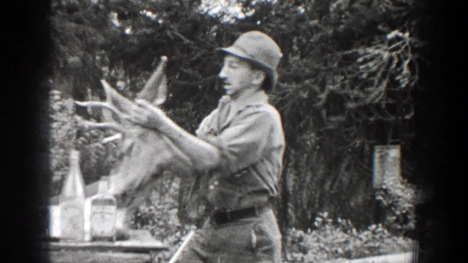 A man in late-1930s Chelan, Washington footage steadies a deer head with antlers near bottles on a table, with trees and brush behind him.