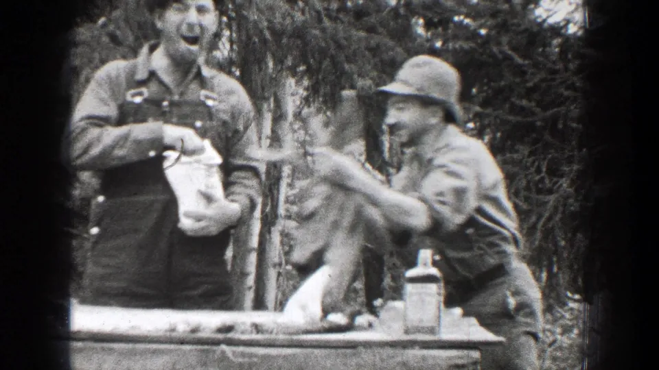 Two adult men appear in the Chelan, Washington home movie as one man holds a buck head near a table while another stands beside him, reacting to the moment.
