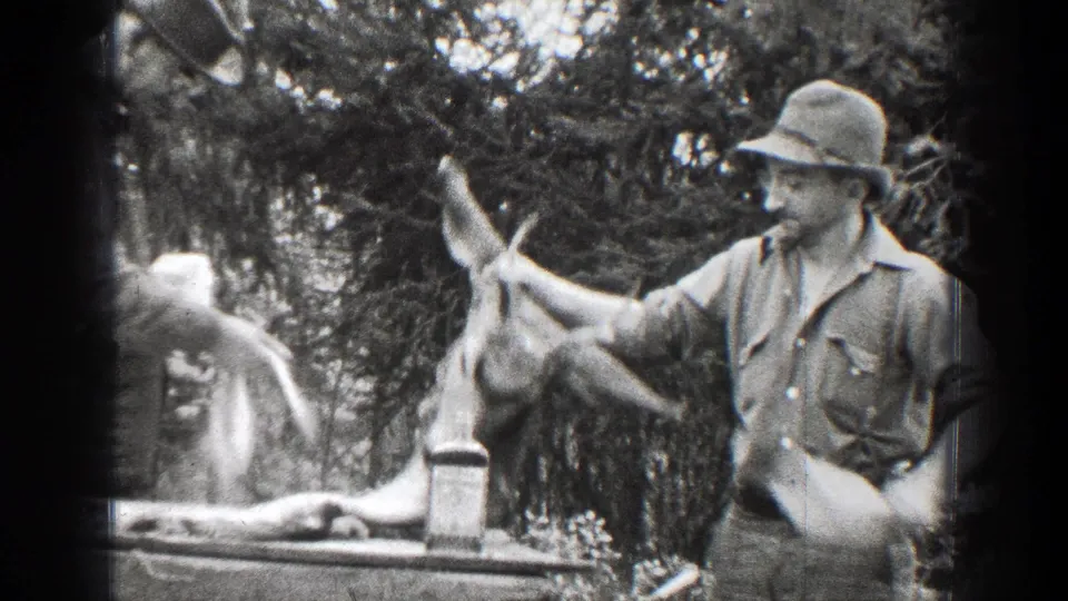 A man in a brimmed hat and shirt holds an antlered deer head at the edge of the frame in 1939 Chelan, Washington outdoor home-movie footage.