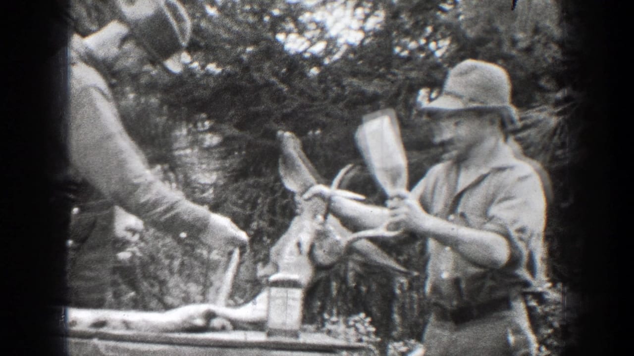 Man in hat and overalls stands beside a deer carcass with antlers in wooded surroundings.