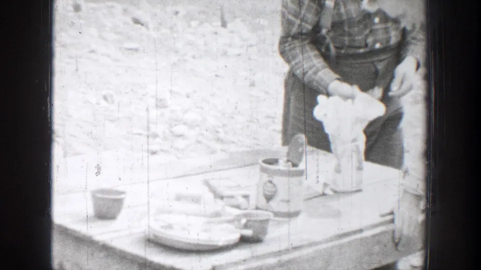 A man in a plaid shirt stands at a small outdoor table with cups, plates, and a pale object in the Chelan home-movie footage.