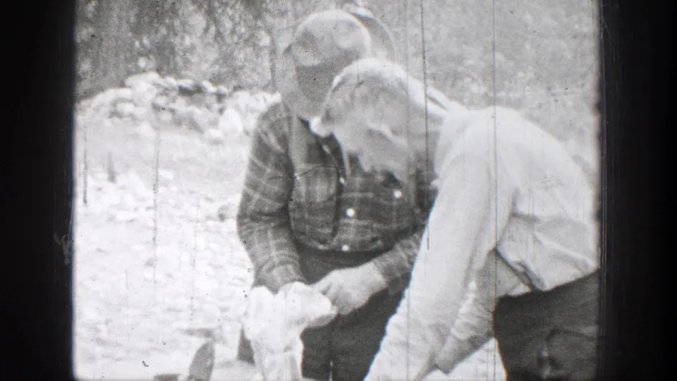 Two men in period outdoor clothing stand together in Chelan as one appears to handle or inspect a light-colored object.