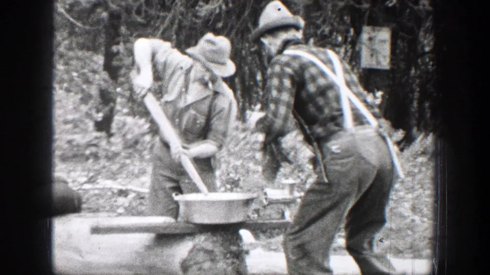 1939 Chelan, Washington frame with two men in hats preparing food over a basin on a rough table in the woods.