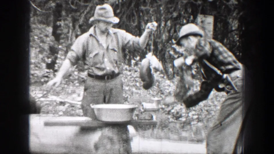 In 1939 Chelan, Washington footage, two men stand by a log table as one holds game meat near a hand grinder and metal pan.