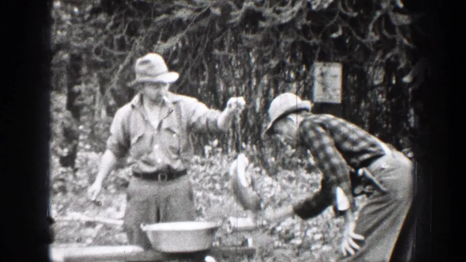 A 1939 Chelan, Washington home-movie frame shows one man holding game meat as another bends near the grinder in a forested area.
