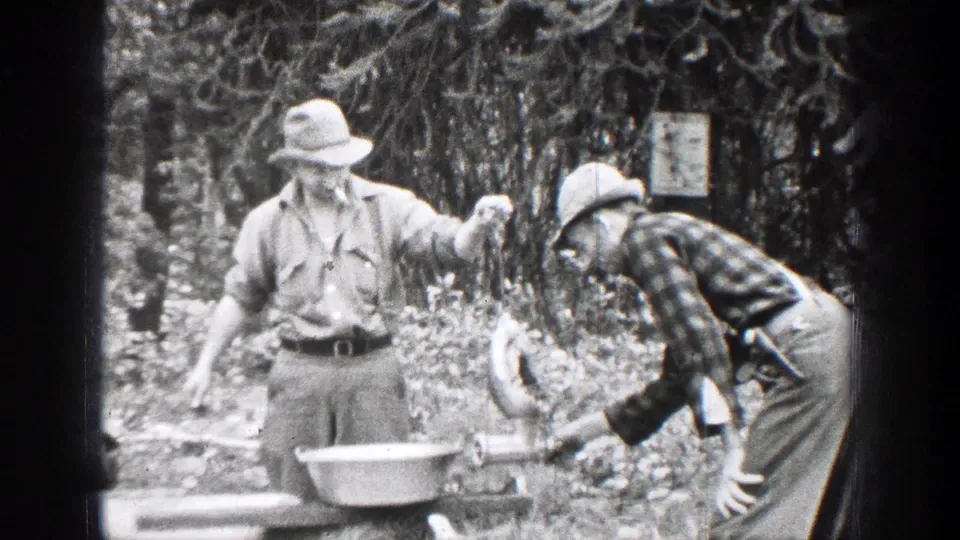 Two men in period outdoor clothing handle game meat beside a hand grinder and metal pan in the Chelan, Washington forest setting.