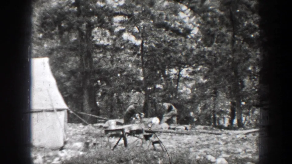 Two men in period outdoor clothing work with logs beside a canvas tent in a rocky forest campsite near Chelan, Washington, in 1939 home-movie footage.