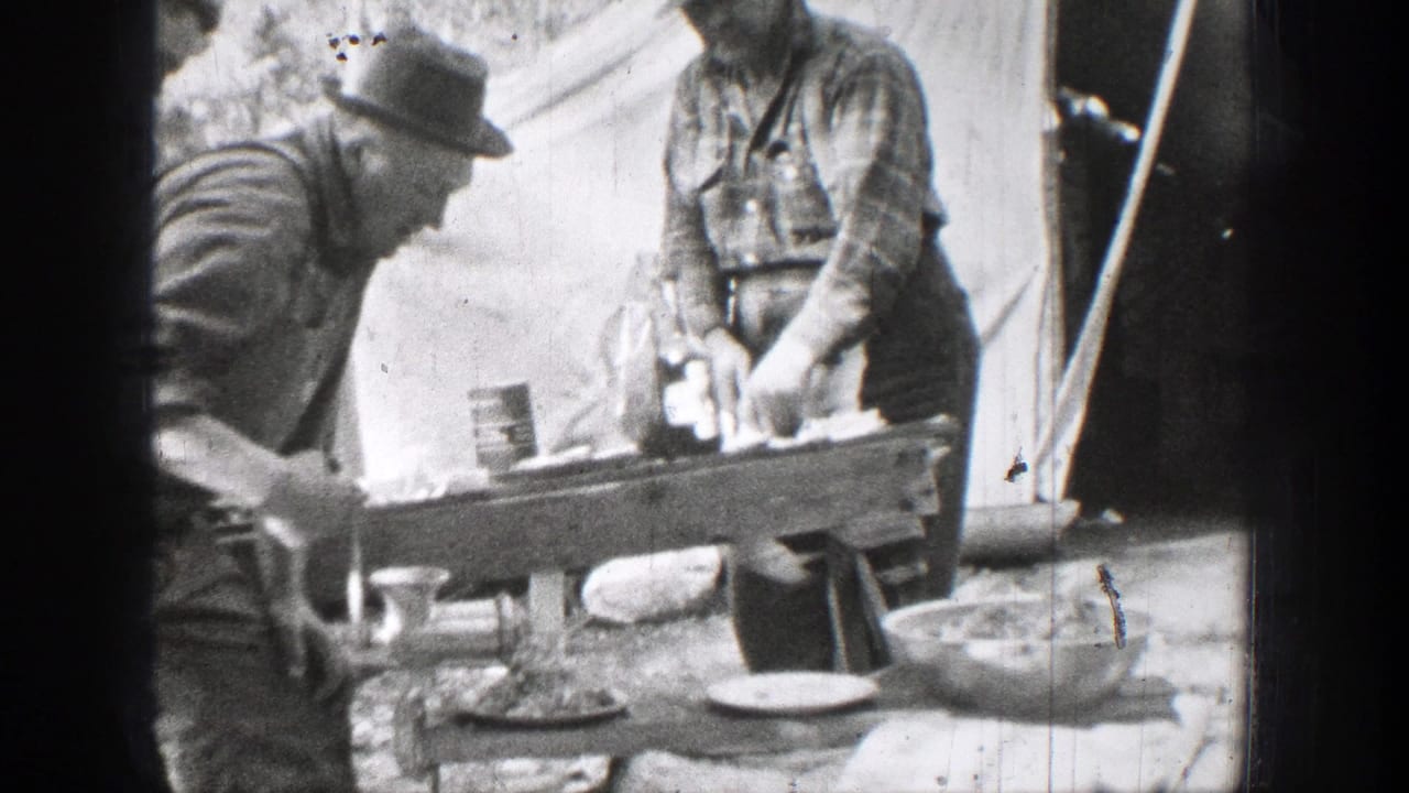 Black-and-white 1939 Chelan, Washington frame showing two men in hats and work clothes preparing food at a wooden table beside a canvas tent or shelter.