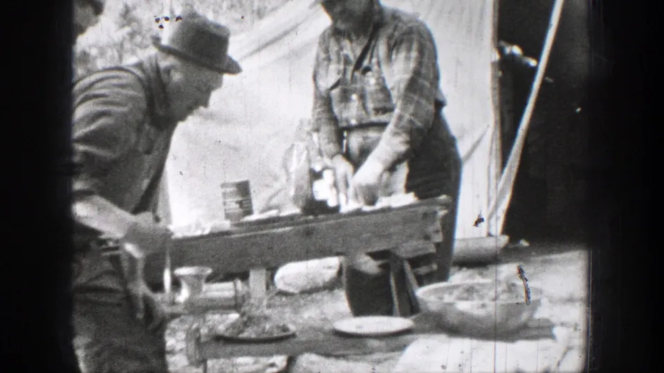 Black-and-white 1939 Chelan, Washington frame showing two men in hats and work clothes preparing food at a wooden table beside a canvas tent or shelter.