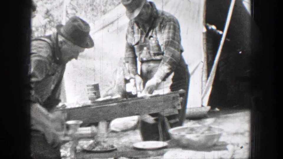 Home-movie frame from 1939 Chelan, Washington, showing men working over a rough outdoor table with cups, bowls, and food preparation items near a canvas shelter.