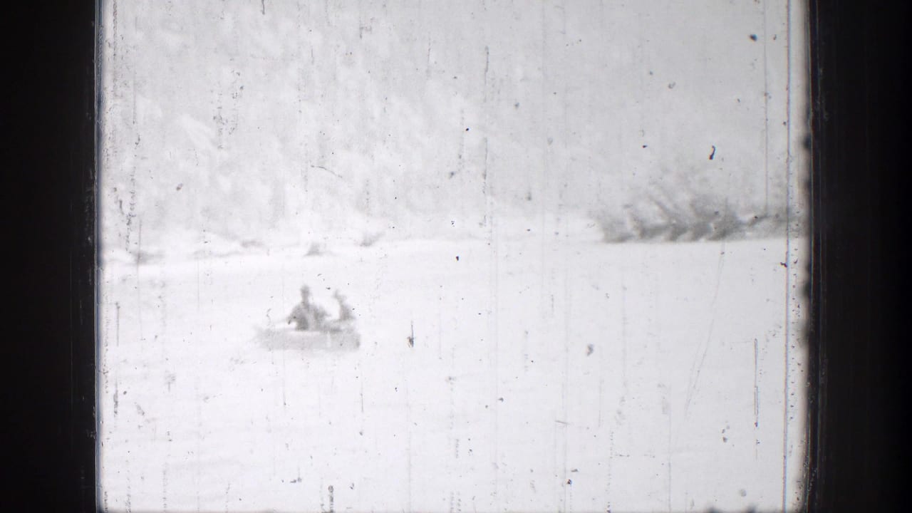 Distant 1939 Chelan, Washington frame of two people in a small boat crossing bright water near a low, brushy shoreline.