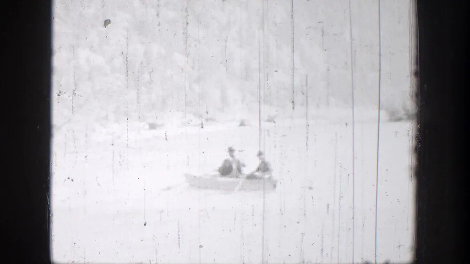 The rowboat remains distant on the Chelan lake, with two people visible through scratched, high-key 8mm footage.