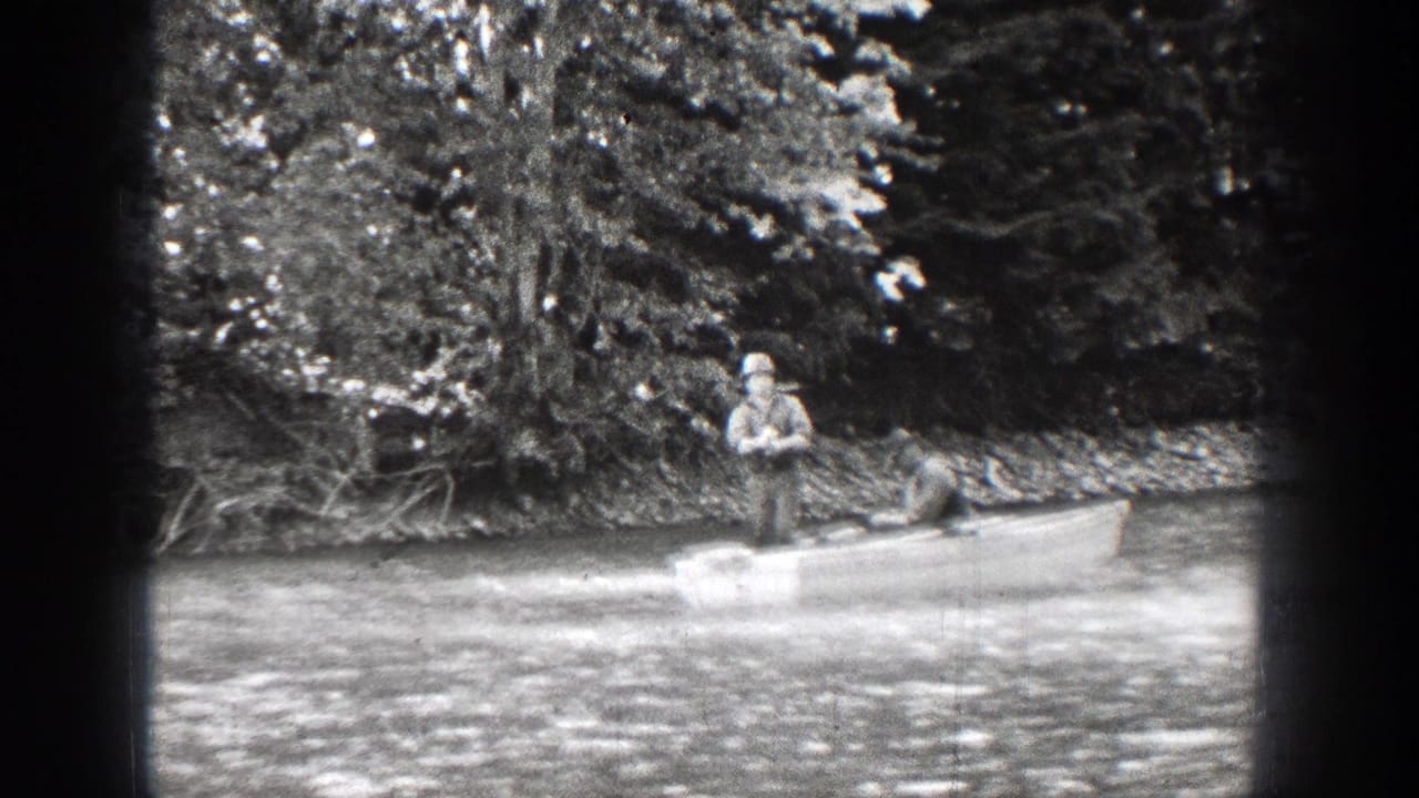 Two men in a small boat near a wooded bank, one standing with fishing gear.