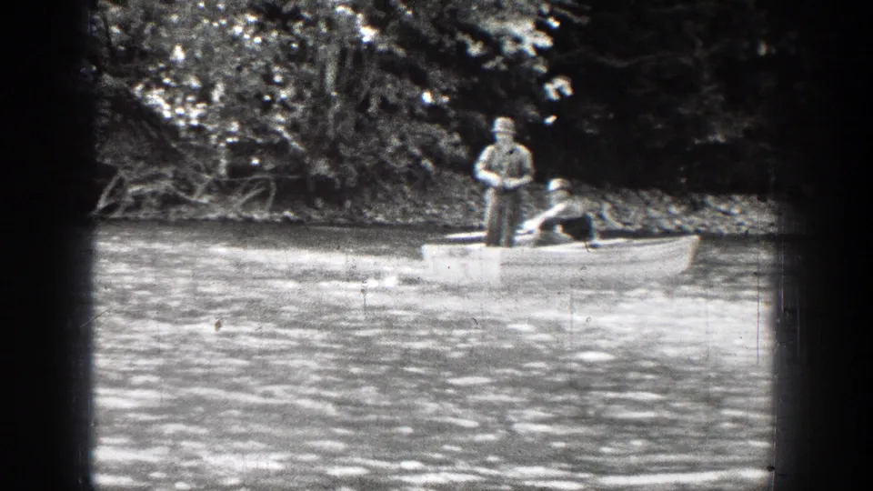 Two men appear in a small rowboat on a Chelan river, with one standing and fishing near a wooded shoreline.