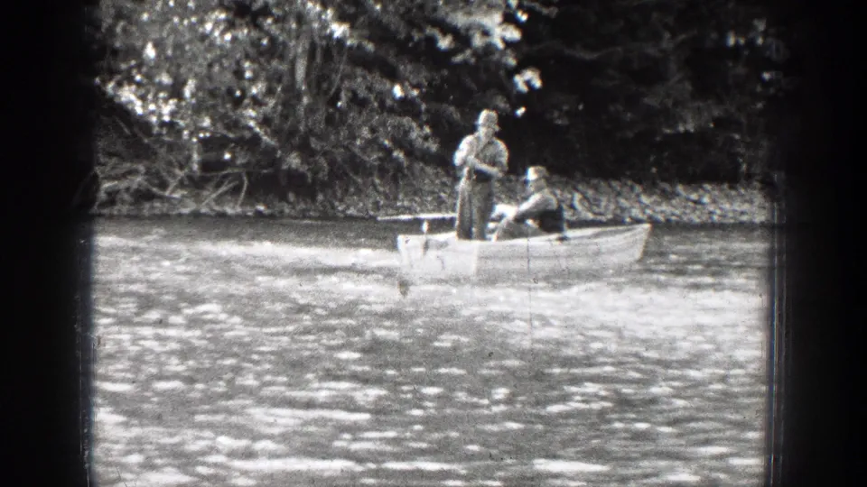 Black-and-white 8mm frame of two adult men in a rowboat on a river in Chelan, Washington, with dense trees behind them.
