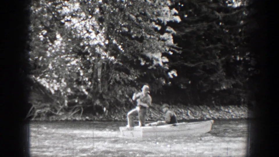 In 1939 Chelan, Washington footage, one man stands at the stern with an oar while another sits in the bow near a wooded riverbank.