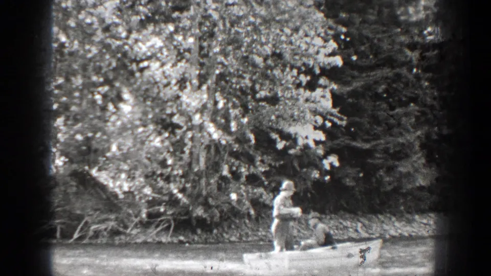 A small rowboat moves past the wooded bank as one man stands with an oar and the other remains seated in the bow.