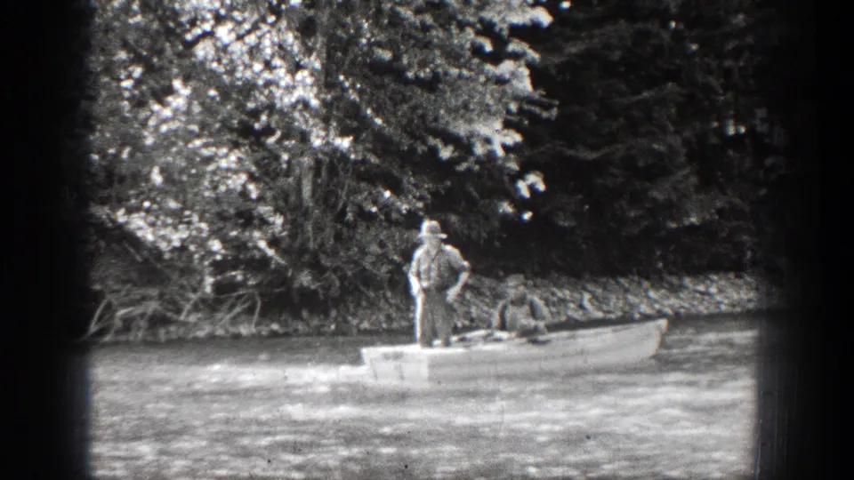 A man stands in the stern of a small boat while another sits at the bow, with a shaded wooded riverbank behind them.