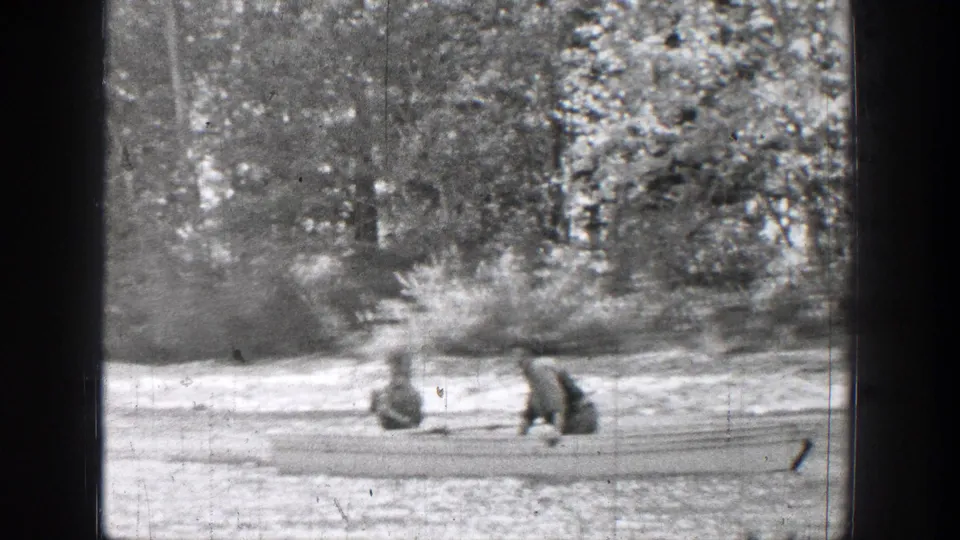 Two distant people in a small boat on Chelan water, with dense trees and a pale shoreline behind them.