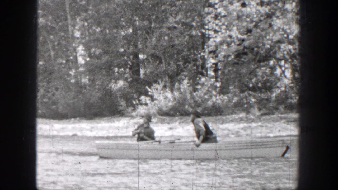 1939 Chelan, Washington home-movie frame showing two people seated in a canoe on a quiet river near a wooded bank.