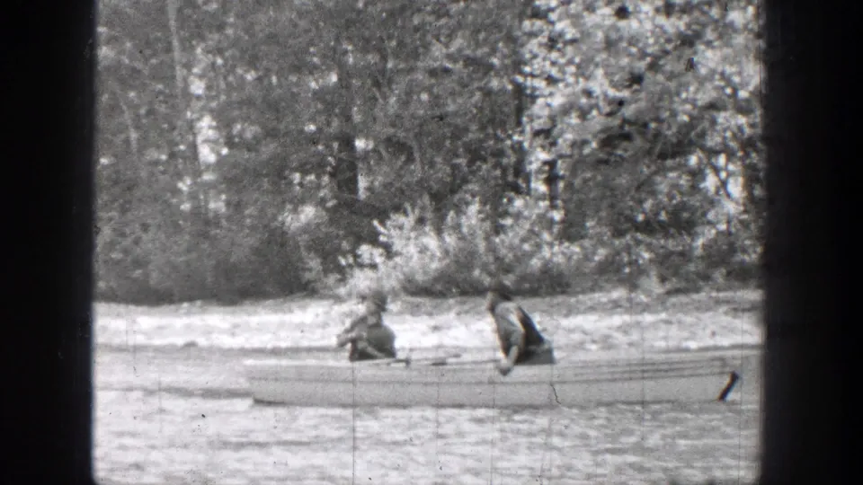 1939 Chelan, Washington home-movie frame showing two people seated in a canoe on a quiet river near a wooded bank.