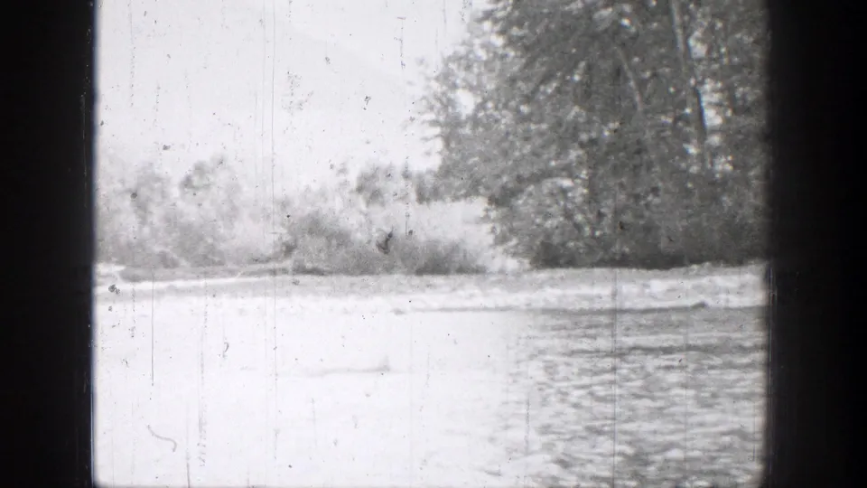 Chelan-area river view in black-and-white 8mm footage, with water, brush, and trees along the far bank.