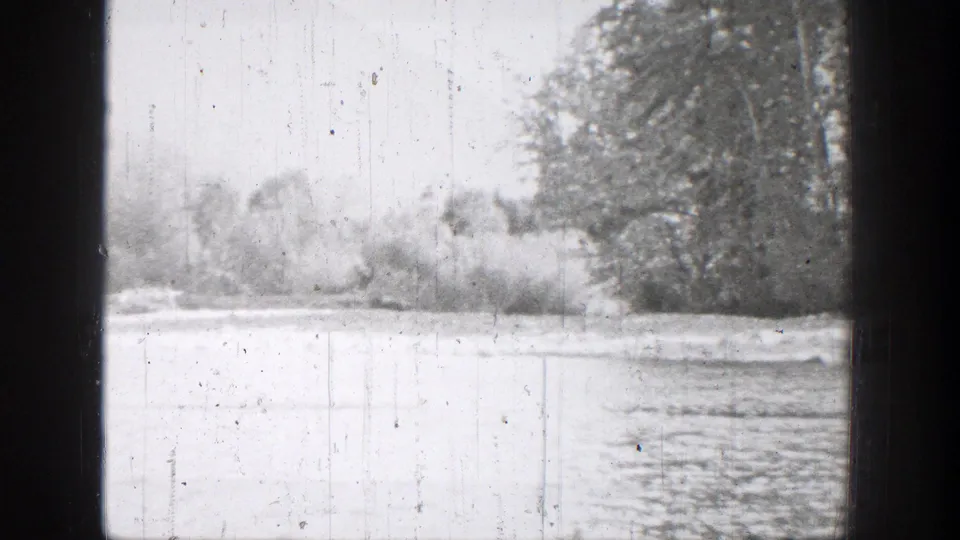 Archival 1939 Chelan river frame showing water, brush, and a tree-lined bank in soft black-and-white 8mm texture.