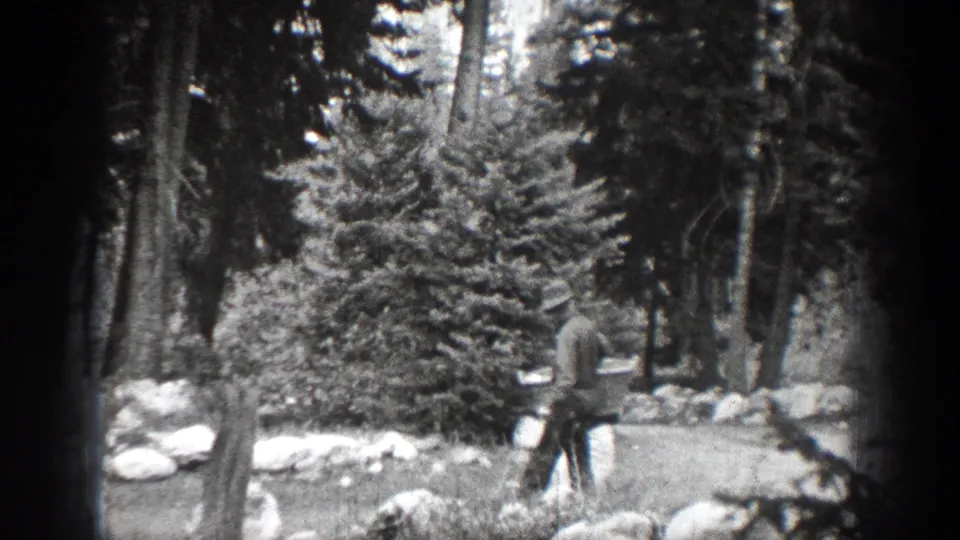 In 1939 Chelan, Washington footage, a man in dark clothing and a hat walks among evergreen trees carrying a light-colored bag.