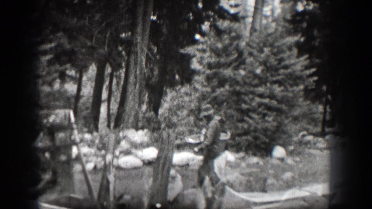 A man in a hat stands among trees in the Chelan-area home movie, near a light-colored hammock or bag-like object.
