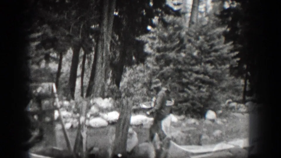 A man in a hat stands among trees in the Chelan-area home movie, near a light-colored hammock or bag-like object.