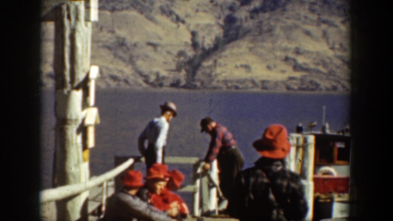 Men in hats and work shirts gather by a dock and small boat on Lake Chelan, with arid mountains rising across the water.