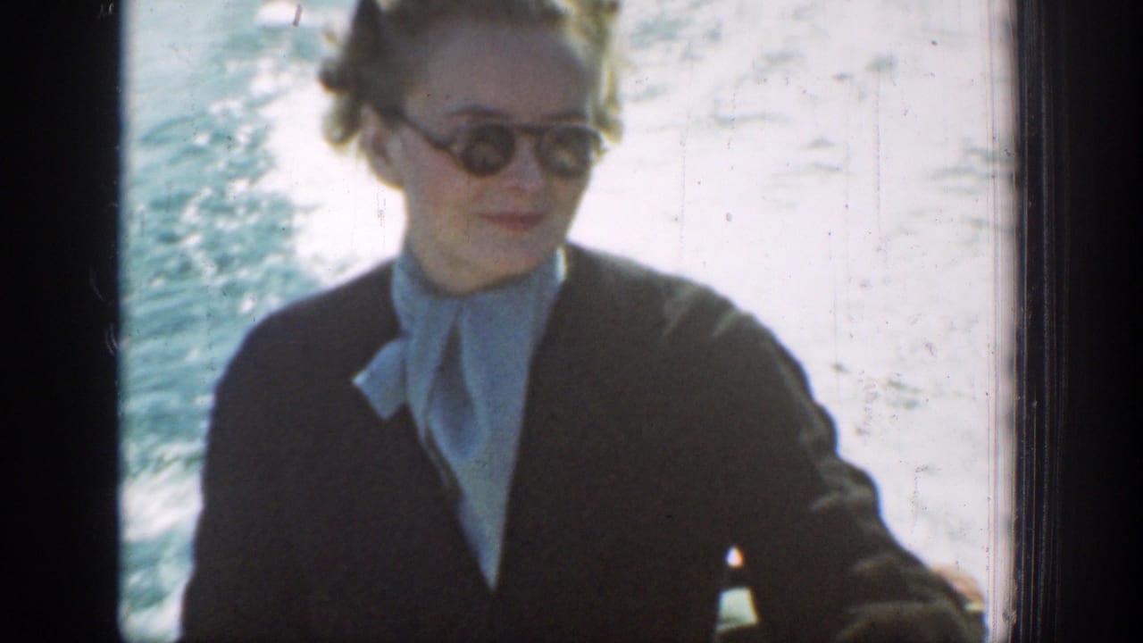 A woman in sunglasses and a dark coat sits on the boat, with bright lake water and wake behind her.