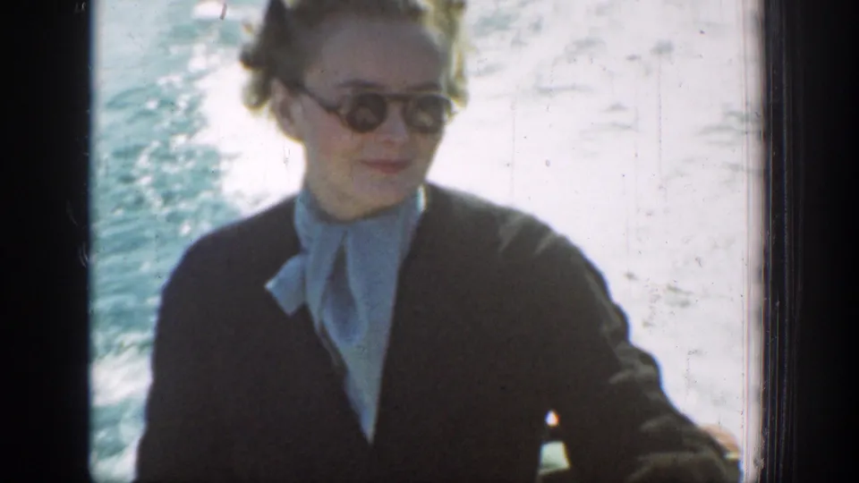 A woman in sunglasses and a dark coat sits on the boat, with bright lake water and wake behind her.