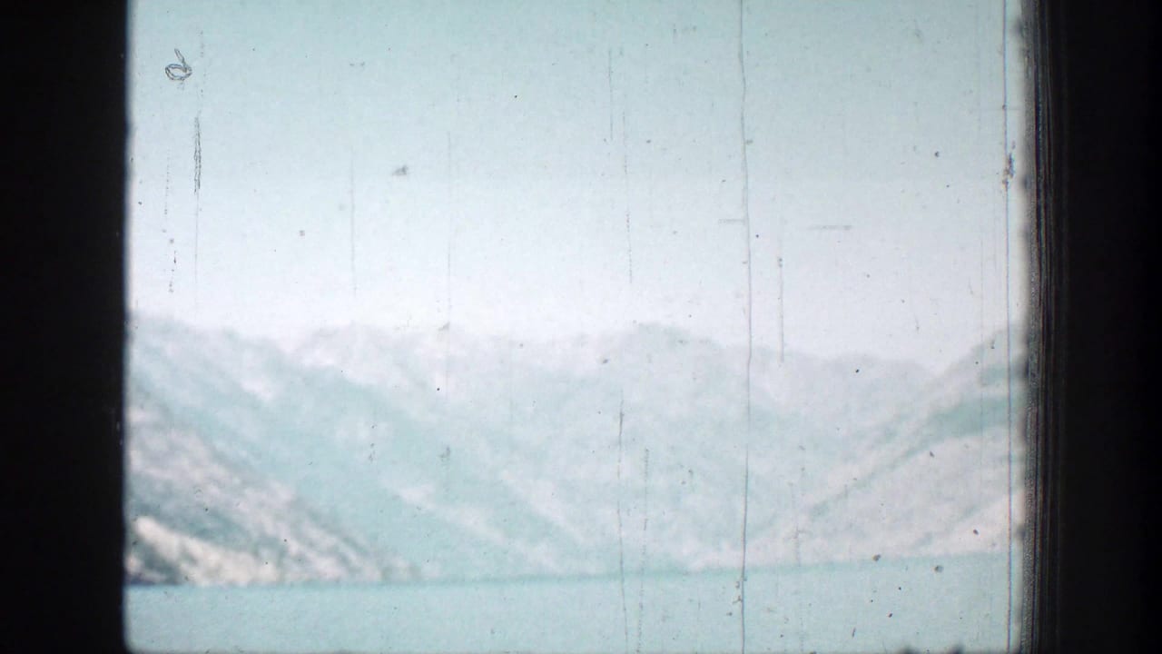 Soft 1939 8mm view of a calm lake and pale mountain ridges in the Chelan, Washington area, partly framed by dark vehicle or window edges.