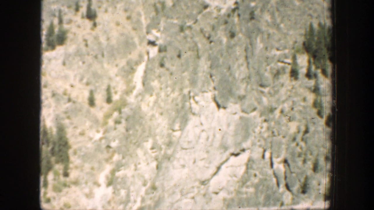 Pale rocky mountain slope with sparse trees and dark film borders.