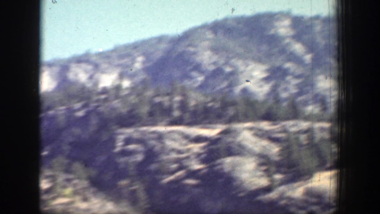 Soft-focus rocky mountain slope with evergreen trees under pale blue sky.