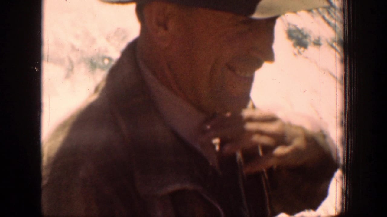 Older man in a brimmed hat and brown jacket smiles beside a window.