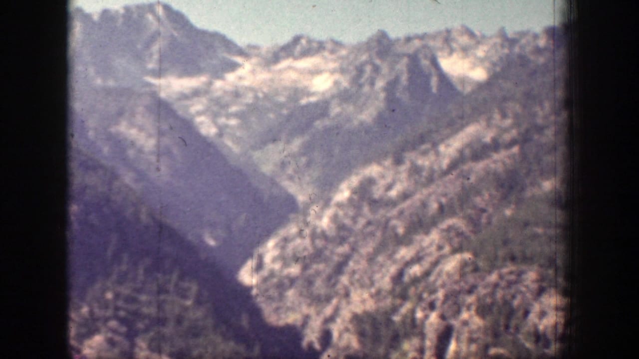 1939 Chelan, Washington home-movie frame showing rugged mountain ridges, forested slopes, and distant snow patches under hazy light.