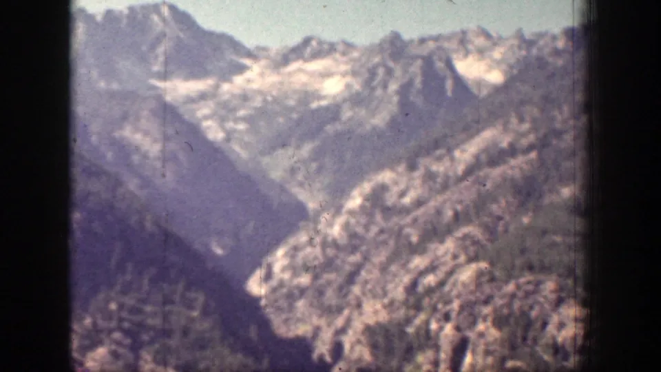 1939 Chelan, Washington home-movie frame showing rugged mountain ridges, forested slopes, and distant snow patches under hazy light.