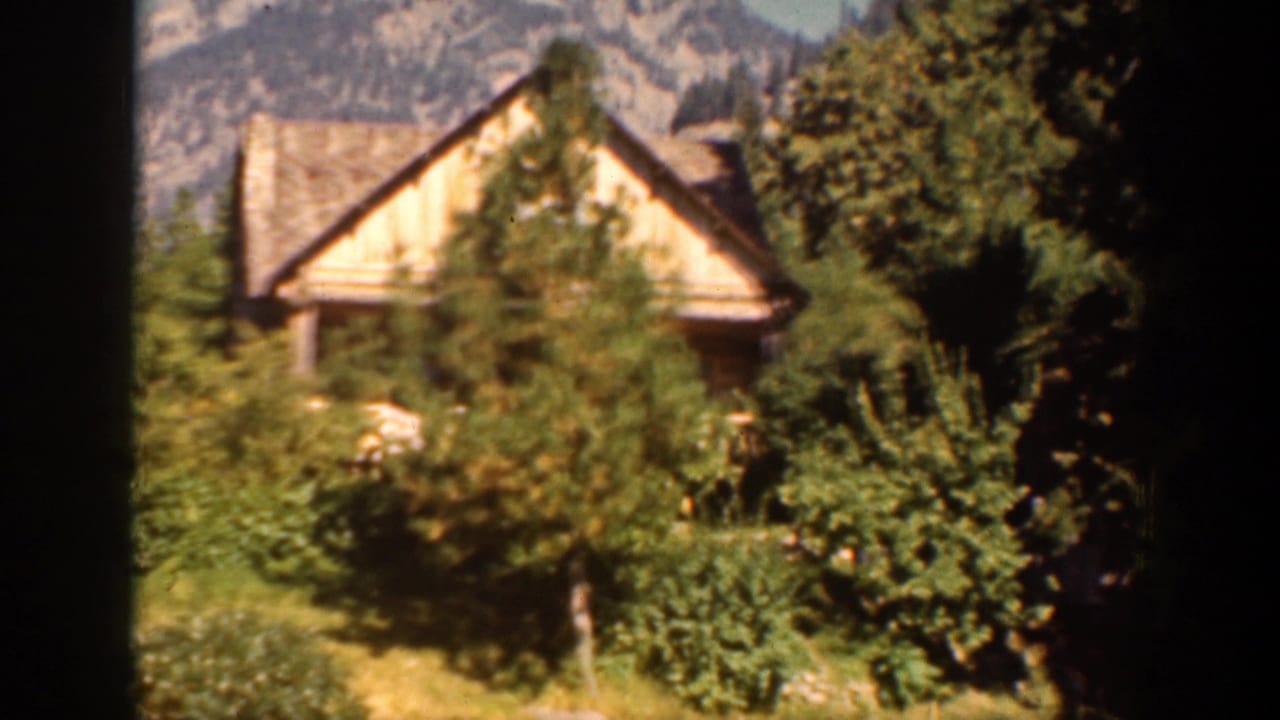1939 Chelan, Washington home-movie frame showing a rustic wooden cabin among trees with rocky mountains in the background.