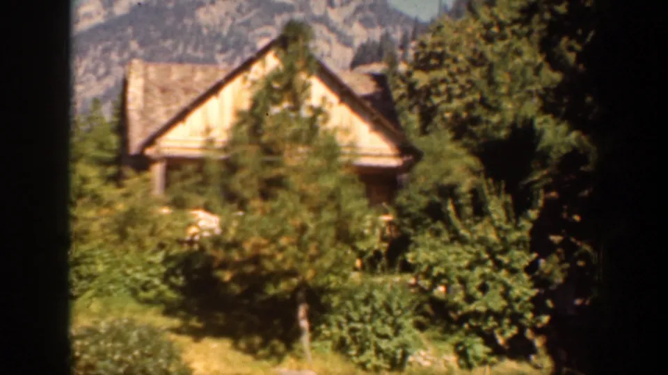 1939 Chelan, Washington home-movie frame showing a rustic wooden cabin among trees with rocky mountains in the background.
