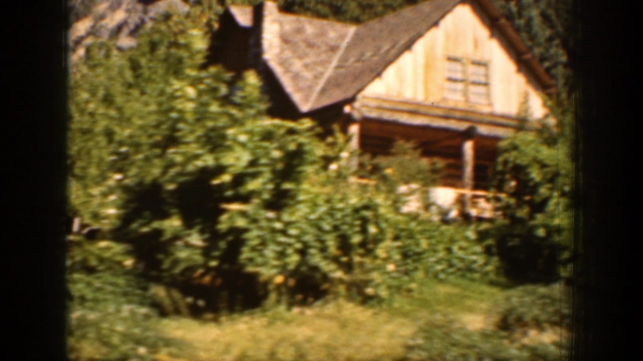 1939 Chelan-area frame of a wood cabin and porch surrounded by thick summer greenery and trees.
