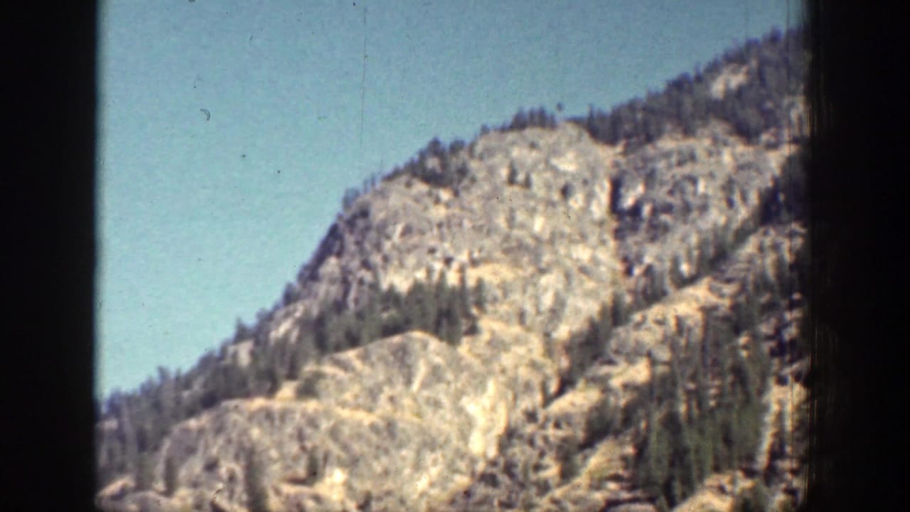 Vintage 8mm view of a steep Chelan-area mountain slope, with pale rock, evergreens, and open blue sky.