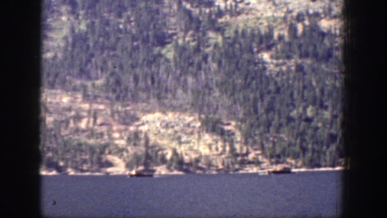 1939 Chelan, Washington lake view showing blue water, a forested mountain shoreline, and small boats in the distance.